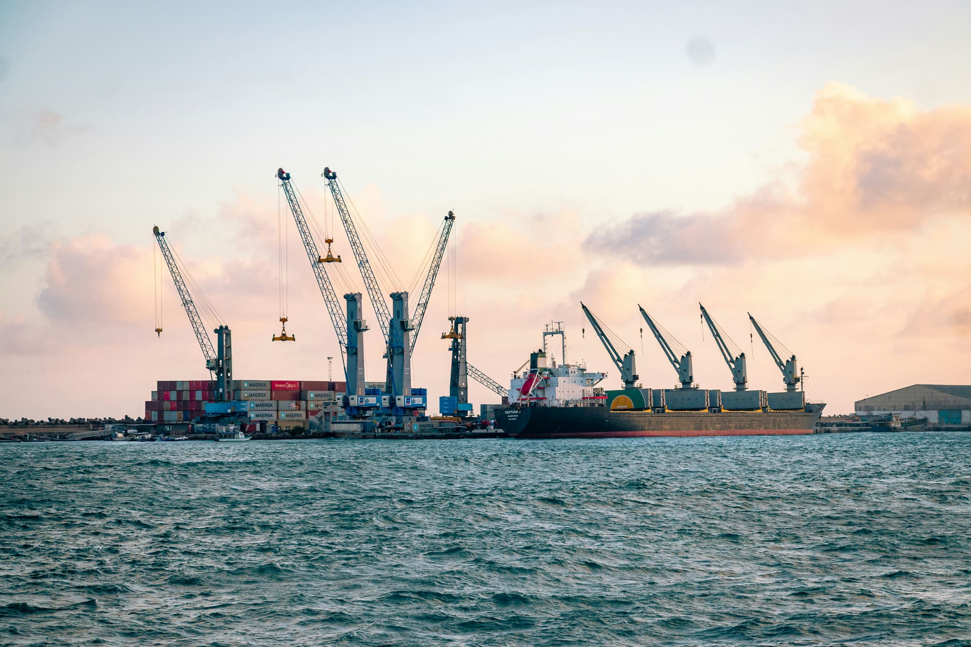 A cargo ship berthed at dusk with its deck cranes raised, shore cranes working containers stacked along the quay behind it, seen from the water.